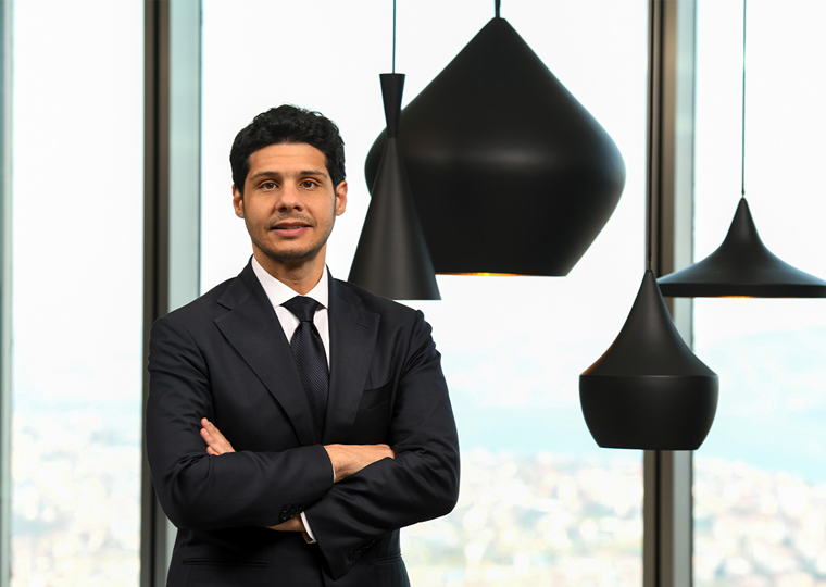 30’larında bir erkek, siyah takım elbise giymiş. Koyu renk bir kravat takıyor. Kollarını göğsünde birleştirmiş şekilde duruyor. Arka planda büyük cam pencerelerden gün ışığı geliyor ve siyah sarkıt lambalar bulunuyor.