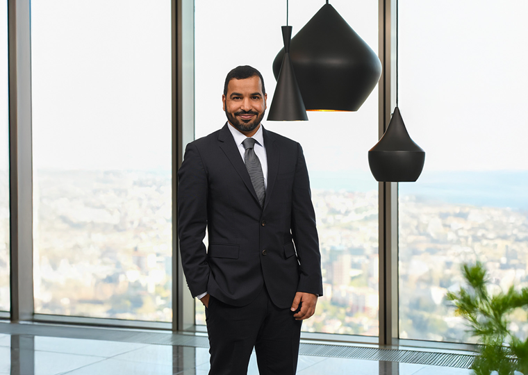 30’larında bir erkek, koyu renk takım elbise giymiş. Gri bir kravat takıyor. Bir eli cebinde, gülümseyerek kameraya bakıyor. Arka planda büyük cam pencereler ve modern siyah sarkıt lambalar bulunuyor.