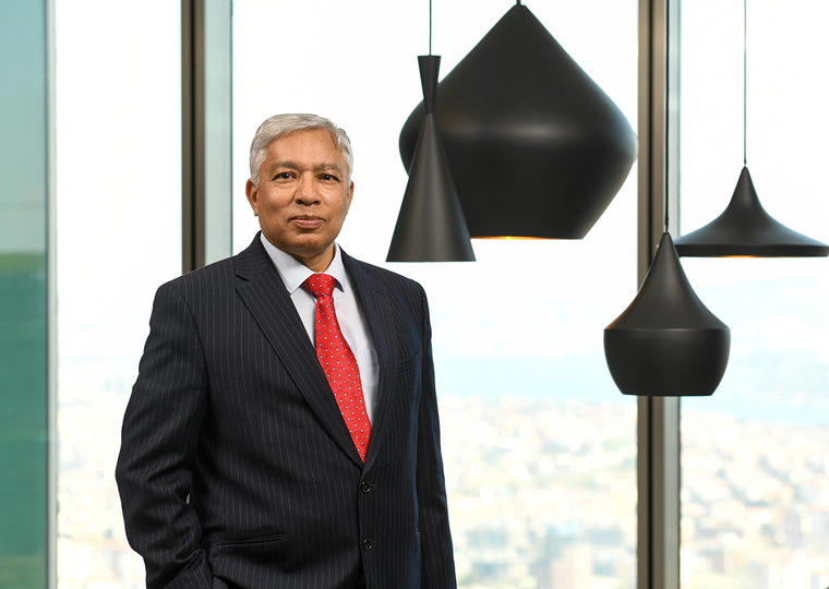 50’lerinde bir erkek, koyu renk çizgili takım elbise giymiş. Kırmızı bir kravat takıyor. Ciddi bir ifadeyle kameraya bakıyor. Arka planda büyük cam pencereler ve modern siyah sarkıt lambalar var.