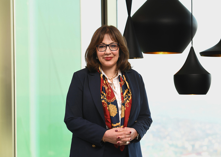 40’larında bir kadın, lacivert ceket ve beyaz gömlek giymiş. Boynunda kırmızı desenli bir fular var. Bordo gözlük takıyor ve gülümseyerek kameraya bakıyor. Arka planda büyük cam pencereler ve modern siyah sarkıt lambalar bulunuyor.