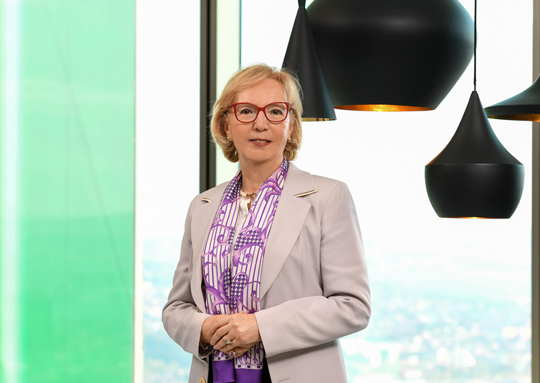 50’lerinde bir kadın, açık renk bir ceket giymiş. Desenli bir fular takıyor. Kırmızı gözlük takmış ve gülümseyerek kameraya bakıyor. Arka planda büyük cam pencereler ve modern siyah sarkıt lambalar yer alıyor.