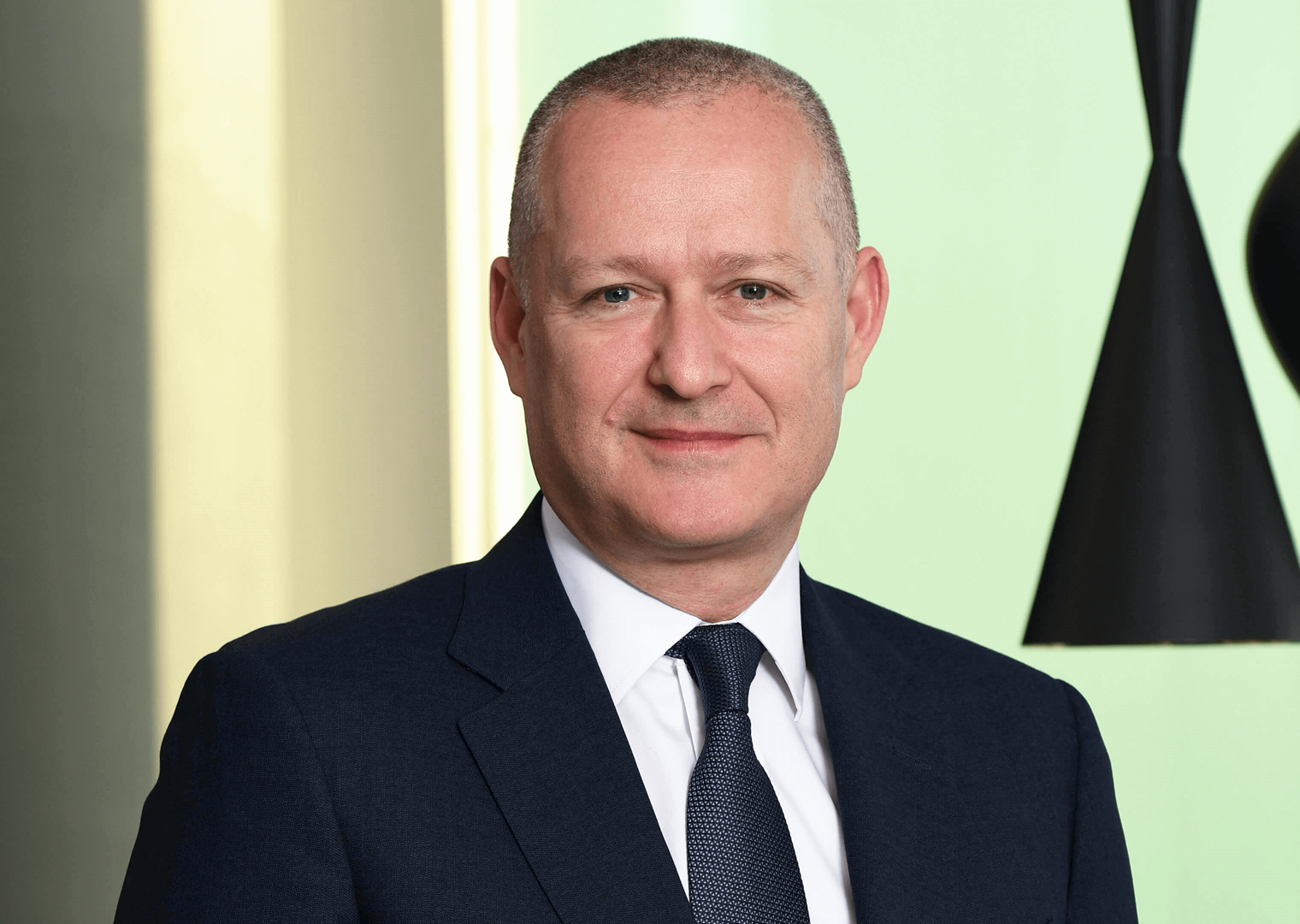 40’larında bir erkek, koyu lacivert takım elbise giymiş. Koyu renk bir kravat takıyor. Gülümseyerek kameraya bakıyor. Arka planda açık renk bir duvar ve siyah sarkıt lambalar var.