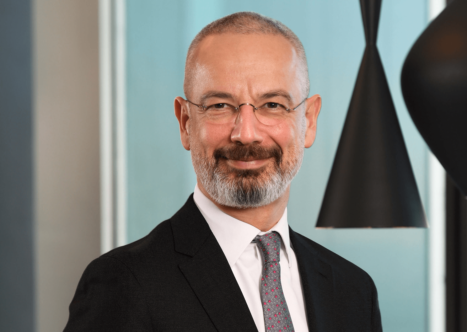 40’larında bir erkek, siyah takım elbise giymiş. Gri desenli bir kravat takıyor. Gözlük takmış ve gülümseyerek kameraya bakıyor. Arka planda açık renk bir duvar ve siyah sarkıt lambalar yer alıyor.