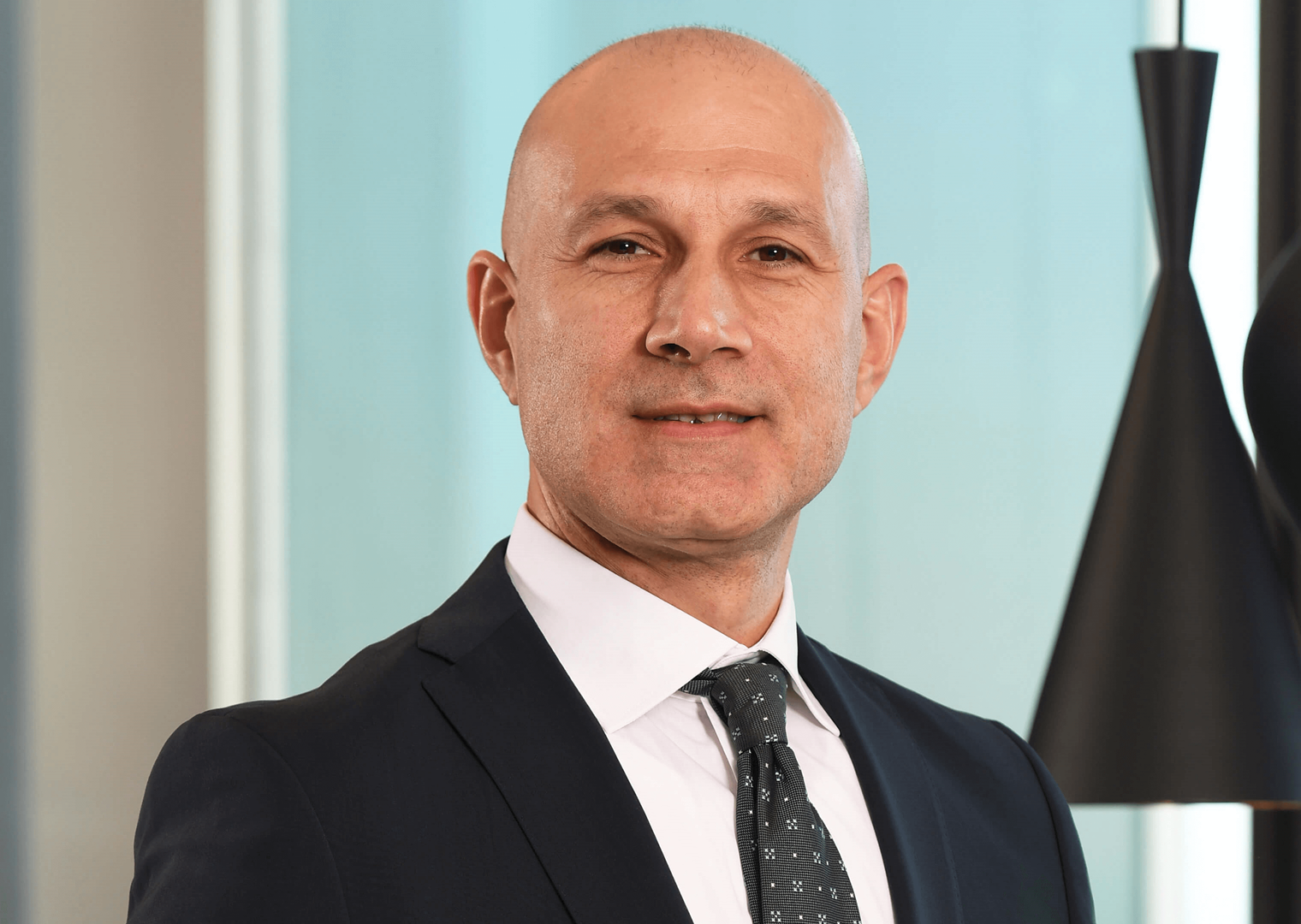 40’larında bir erkek, koyu renk takım elbise giymiş. Desenli bir kravat takıyor. Gülümseyerek kameraya bakıyor. Arka planda açık renk bir duvar ve siyah sarkıt lambalar bulunuyor.