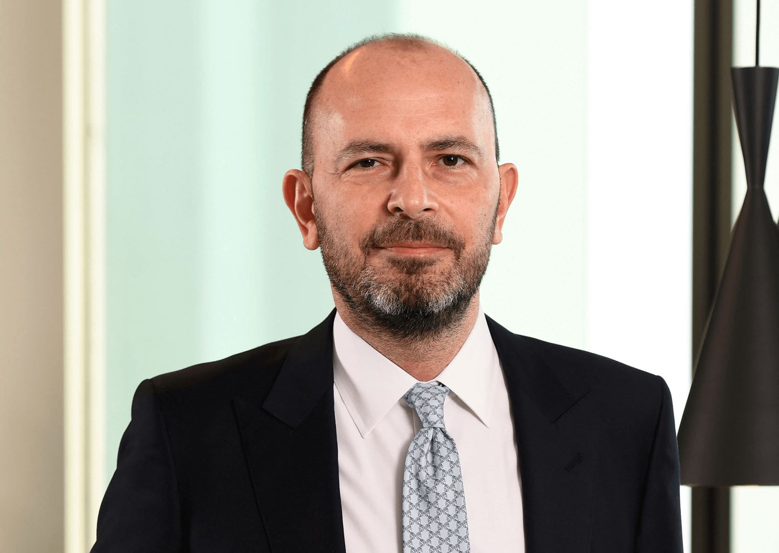 40’larında bir erkek, lacivert takım elbise giymiş. Açık mavi bir kravat takıyor. Ciddi bir ifadeyle kameraya bakıyor. Arka planda açık renk bir duvar ve siyah sarkıt lambalar yer alıyor.