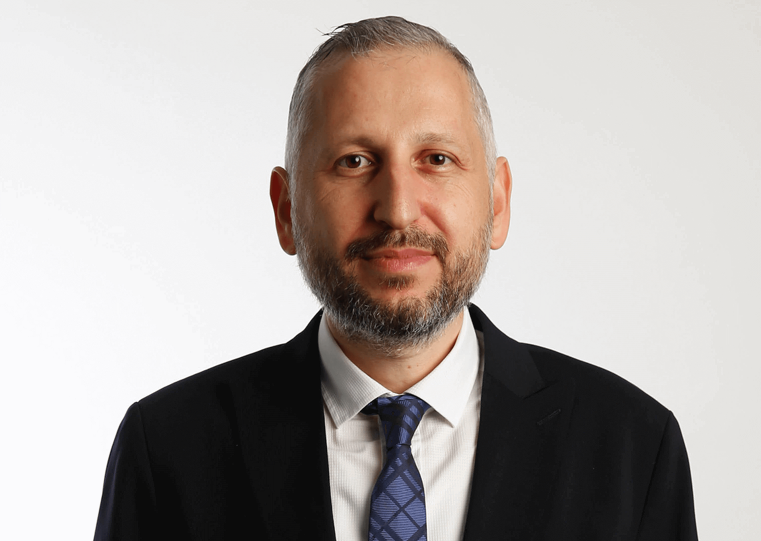 40’larında bir erkek, siyah takım elbise giymiş. Beyaz gömlek ve lacivert desenli bir kravat takıyor. Gülümseyerek kameraya bakıyor. Arka planda beyaz bir zemin bulunuyor.