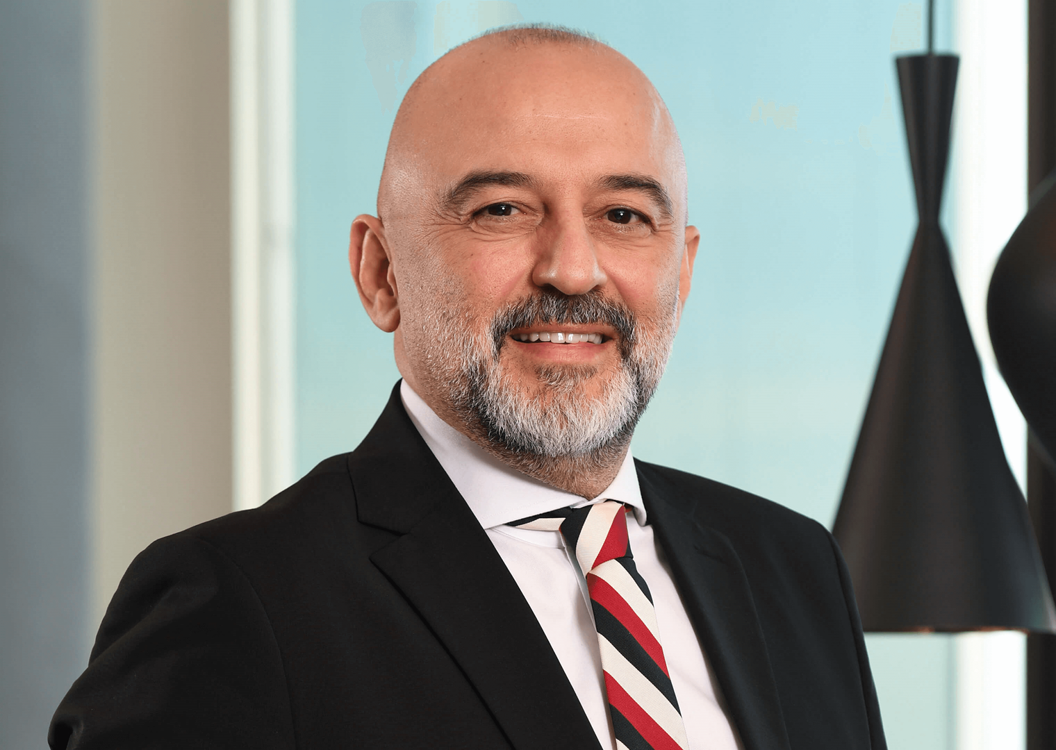 50’lerinde bir erkek, siyah takım elbise giymiş. Kırmızı, siyah ve beyaz çizgili bir kravat takıyor. Gülümseyerek kameraya bakıyor. Arka planda modern siyah sarkıt lambalar bulunuyor.