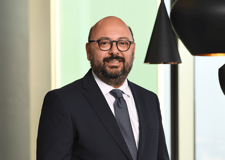 40’larında bir erkek, lacivert takım elbise giymiş. Gri desenli bir kravat takıyor. Gülümseyerek kameraya bakıyor. Arka planda modern siyah sarkıt lambalar yer alıyor.