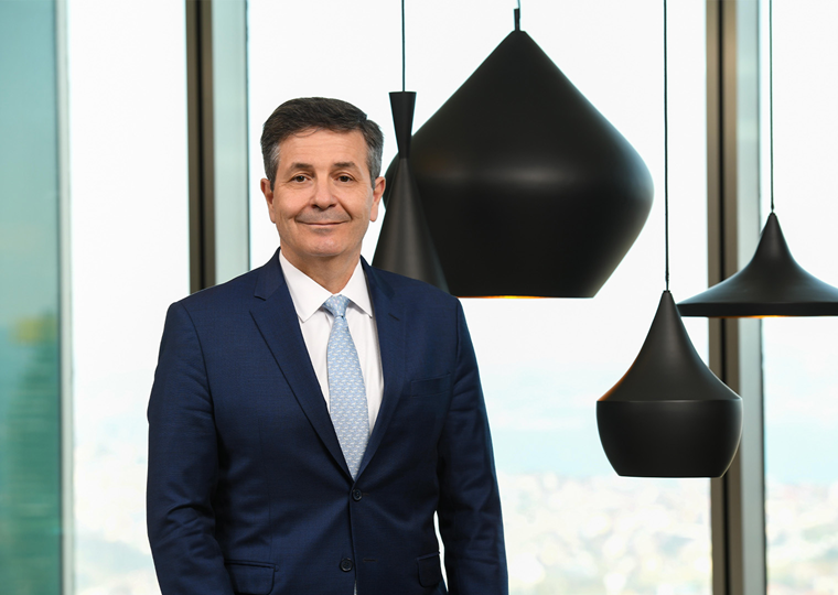 50'lerinde bir erkek. Lacivert takım elbise giymiş. Açık mavi kravat takıyor. Gülümseyerek kameraya bakıyor. Arka planda büyük cam pencereler ve modern siyah sarkıt lambalar var.