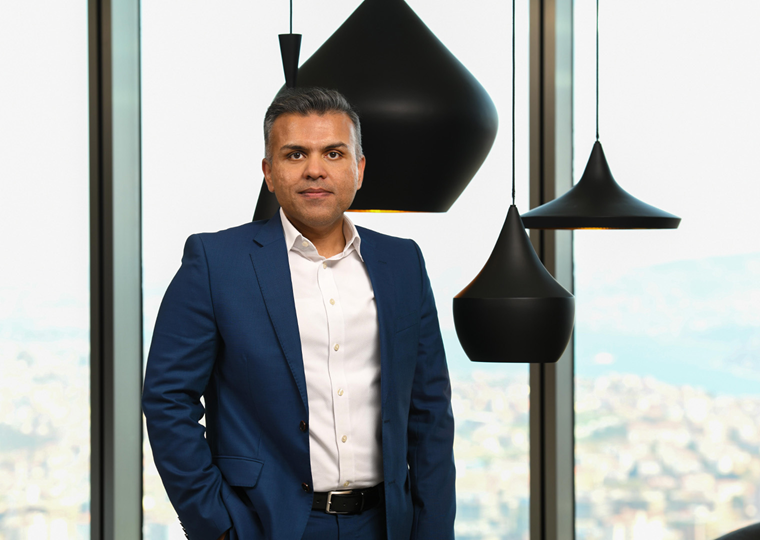 40’larında bir erkek, beyaz gömlek ve lacivert takım elbise giymiş. Bir eli cebinde duruyor. Arka planda şehir manzaralı büyük cam pencereler ve siyah sarkıt lambalar var.