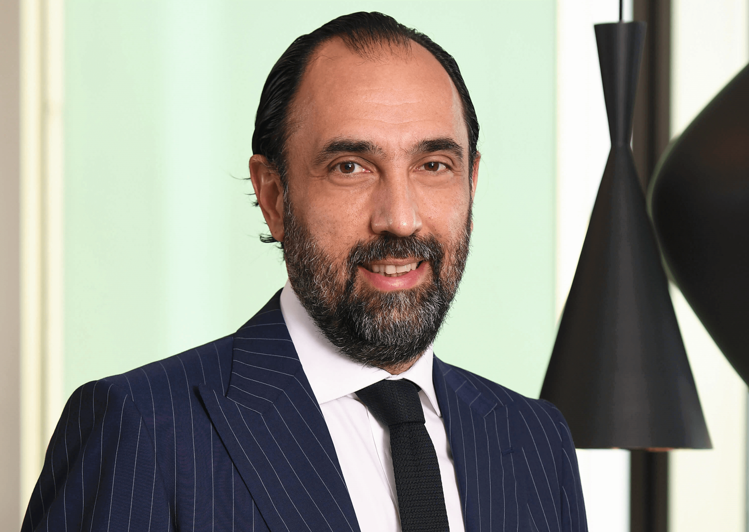 40’larında bir erkek lacivert çizgili takım elbise giymiş. Koyu renk bir kravat takıyor. Gülümseyerek kameraya bakıyor. Arka planda açık renk bir duvar ve modern siyah sarkıt lambalar bulunuyor.