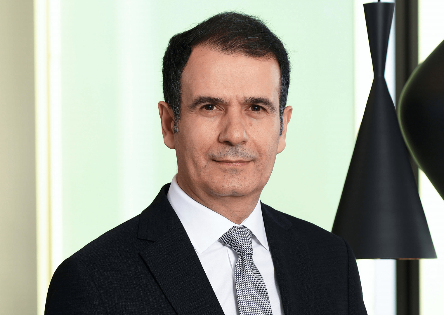 50’lerinde bir erkek siyah takım elbise giymiş. Gri tonlarında bir kravat takıyor. Ciddi bir ifadeyle kameraya bakıyor. Arka planda açık renk bir duvar ve siyah sarkıt lambalar var.