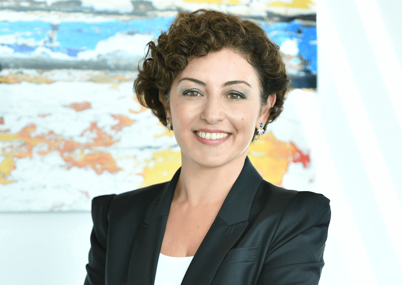 40’larında bir kadın siyah ceket ve beyaz bluz giymiş. Gülümseyerek kameraya bakıyor. Arka planda renkli bir tablo yer alıyor.
