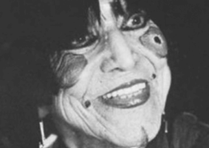 Siyah beyaz bir portrede, yüzü detaylı şekilde boyanmış bir kişi bulunuyor. Göz çevresinde koyu halkalar, yanaklarında desenler var. Saçları siyah ve alnına düşüyor, dudaklarında belirgin bir gülümseme yer alıyor.