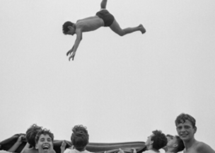 Açık gri arka plan önünde, ortada havada yüzüstü şekilde duran genç bir erkek var, altında kahkaha atan ve bir örtü tutan altı genç yer alıyor, neşeli bir an canlandırılmış.