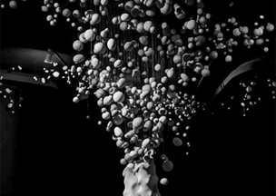 Siyah arka planda, tavandan sarkan iplerde farklı boyutlarda çok sayıda yuvarlak nesne asılı, aşağıda dalgalı biçimli büyük beyaz bir kütle bulunuyor. Yukarıya doğru yükselen bu beyaz formlar, bir patlama ya da enerji akışı etkisi yaratıyor