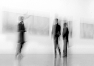 Bir galeride gezenl üç kişinin siyah beyaz ve bulanık fotoğrafı. Duvarlarda yatay çerçeveli tablolar asılı.