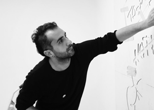 Siyah beyaz fotoğrafta 30’larında kısa saçlı bir erkek beyaz duvara çizim yapıyor. Koyu renk uzun kollu bir kazak giyiyor.