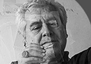 60’larında beyaz saçlı bir erkek elini kaldırmış şekilde elinde bulunan nesneye bakıyor. Üzerinde koyu renk bir kıyafet var. Görsel siyah beyaz tonlarda.