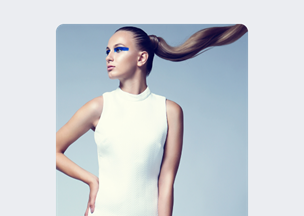 Gri arka plan önünde, 20’lerinde bir kadın belden yukarısı kadrajda duruyor, beyaz bir elbise giymiş, göz çevresinde kalın mavi bir makyaj hattı var, uzun at kuyruğu saçları arkaya savrulmuş.