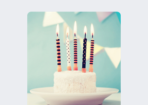 Açık mavi arka plan önünde beyaz kremalı küçük bir pasta duruyor, üzerinde yanmakta olan beş adet mum bulunuyor, arka planda pastel tonlarda üçgen süslemeler yer alıyor.