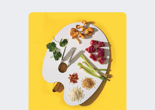 Sarı zemin üzerinde beyaz bir ressam paleti, üzerinde doğranmış kırmızı et parçaları, mantarlar, yeşil soğanlar, kişniş yaprakları, pirinç taneleri, pul biber ve susam yer alıyor, paletin ortasında metal bir maşa bulunuyor.