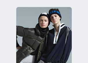 Açık gri arka plan önünde iki kadın yan yana duruyor, biri koyu yeşil şişme mont ve siyah bere takmış, diğeri lacivert beyaz çizgili fermuarlı üst giymiş, her ikisinin de yüz ifadesi ciddi.