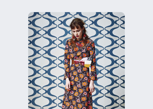 Mavi ve beyaz dalgalı desenli bir duvar önünde, desenli elbise giymiş bir kadın duruyor. Elbisesinde turuncu çiçekler ve siyah çizgiler yer alıyor, belinde büyük bir kurdele detayı bulunuyor.
