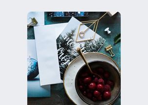 Koyu zemin üzerinde, beyaz zarflar, yılbaşı temalı bir kart, altın renkli ataçlar ve kırmızı meyvelerle dolu bir kase bulunuyor, yanında altın saplı bir kaşık yer alıyor.