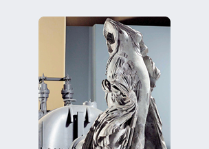 Endüstriyel bir mekânda, pelerinli bir figürü betimleyen taş bir heykel bulunuyor, heykelin yüzeyinde yıpranma izleri var, arka planda metal makineler yer alıyor.