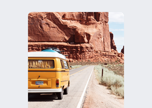 Bir trarafta devasa kırmızı kayalıklarla çevrili bir manzarada, diğer tarafı uçurum. Ortasındaki yolda ilerleyen sarı bir minibüs manzaraya doğru gidiyor, araç üzerinde mavi bir sörf tahtası yer alıyor.