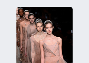 Podyum üzerinde arka arkaya yürüyen bir grup kadın model bulunuyor, her biri ten rengine yakın şeffaf ve ışıltılı elbiseler giymiş, sahne loş bir ışıkla aydınlatılmış.