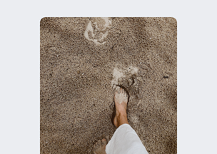 Kumluk zeminde, beyaz pantolon giymiş bir kişi yalınayak yürüyor, ayak izleri kumda belirgin şekilde kalmış.