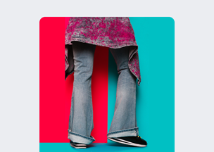 Kırmızı ve mavi zemin önünde, paçaları geniş mavi kot pantolon giyen bir kişi duruyor, ayaklarında siyah spor ayakkabıları, üzerinde mor ve gri tonlarda asimetrik kesimli bir üst var.