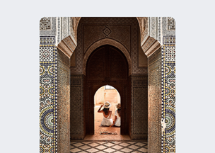 Geometrik süslemelerle bezenmiş kemer biçimli giriş görülüyor, duvarlarda mozaik ve kabartma desenler yer alıyor, kemerin altında oturan iki kişi arkadan fotograflanmış.