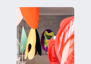 Geniş ve aydınlık bir iç mekânda tavandan sarkıtılmış büyük, renkli formlar yer alıyor, yüzeylerinde kırmızı, turuncu, sarı, yeşil ve mor tonlar hâkim.